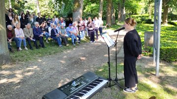 Eine junge Frau steht rechts im Bild, die Hände hinter dem Rücken verschränkt, und spricht mit einem Mikrofon zu einer Gruppe links im Bild.