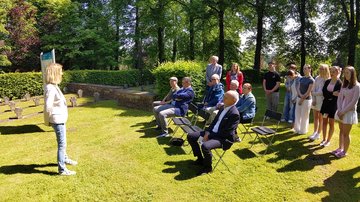 Links im Bild eine Frau mit blonden Haaren, sie hat die Arme auf dem Rücken verschränkt und spricht zu einer Gruppe rechts im Bild die teilweise auf Stühlen sitzen. Im Bildhintergrund eine Tafel und Grabkreuze.