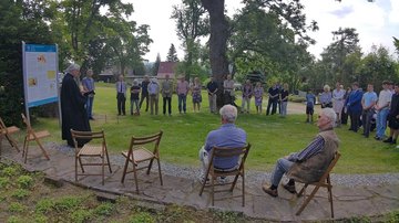 Links im Bild die Tafel, davor der Pastor. Im hHalbkreis dahinter eine Gruppe Menschen die zuhören. Im Vordergrund Stühle, auf zwei von ihnen sitzen Männer mit dem Rücken zum Betrachter des Fotos.