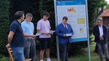 Zwei Männer und drei Schüler stehen links und rechts der Tafel. Die Schüler halten Blätter in den Händen, einer liest von seinem Zettel vor. 
