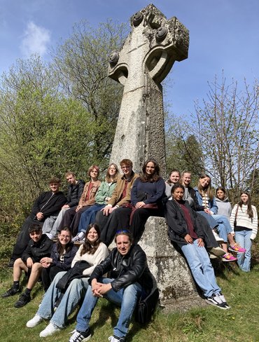Eine Gruppe von Jugendlichen sitzt und steht um das Hochkreuz der Kriegsgräberstätte