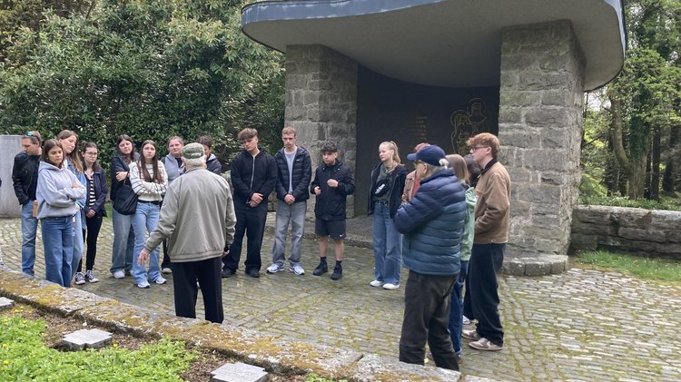 Schülerinnen und Schüler stehen vor einer kleinen Kapelle. Sie hören einem älteren Mannzu, der von hinten zu sehen ist und mit der Hand auf einen Grabstein zeigt.