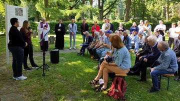 Links spricht ein Mädchen in ein Mikrofon, neben ihr steht ein Junge. Hinter den Beiden eine Tafel mit Text und Abbildungen. Die Menschengruppe rechts hört zu.