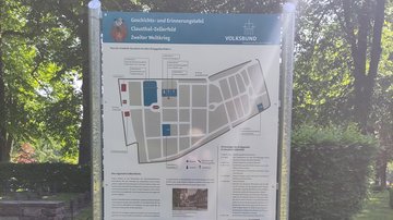 Die Tafel mit einem Friedhofsplan auf dem Gräberfelder der Kriegstoten gekennzeichnet sind. Dazu Texte und weitere Abbildungen. Im Hintergrund leinks eine niedrige Mauer sowie Büsche und Bäume.