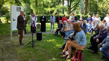 Links spricht ein Mann in das Mikrofon, dahinter eine Tafel mit Text und Abbildungen. Die Menschengruppe rechts hört zu. Im Hintergrund stehen weitere Personen, darunter zwei Pfarrer.