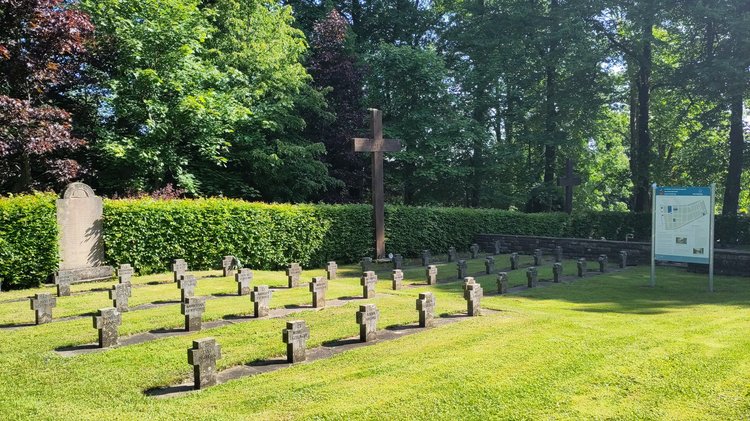 Ein Gräberfeld. Die steinernen Grabkreuze auf einer Rasenfläche, im Hintergrund ein hölzernes Hochreuz. Rechts im Bild die Geschichts- und Erinnerungstafel. Im Hintergrund eine Hecke und Bäume.